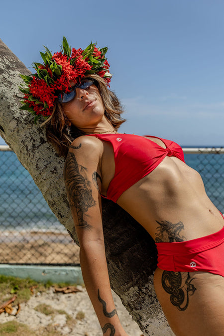 A woman wearing a bright red two-piece bikini and a red floral crown leans against a tree outdoors near the ocean, with a chain-link fence and blue sky in the background.
